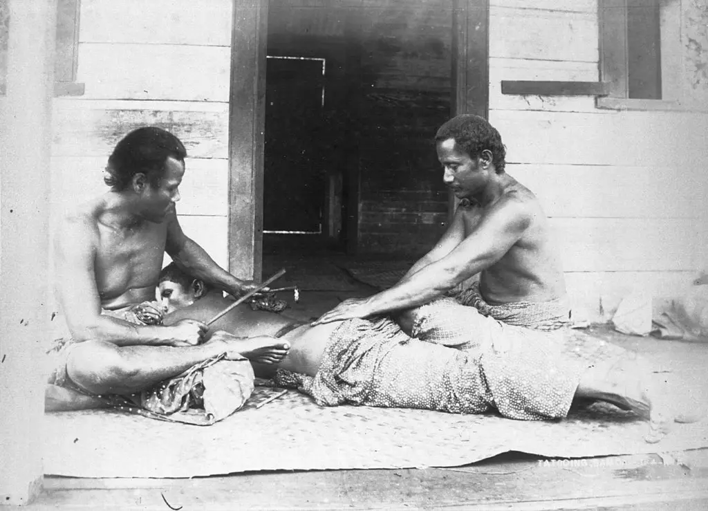A Samoan tattooist and his assistant carry out a traditional tatau using handmade tools, photographed around 1895 by Thomas Andrew.