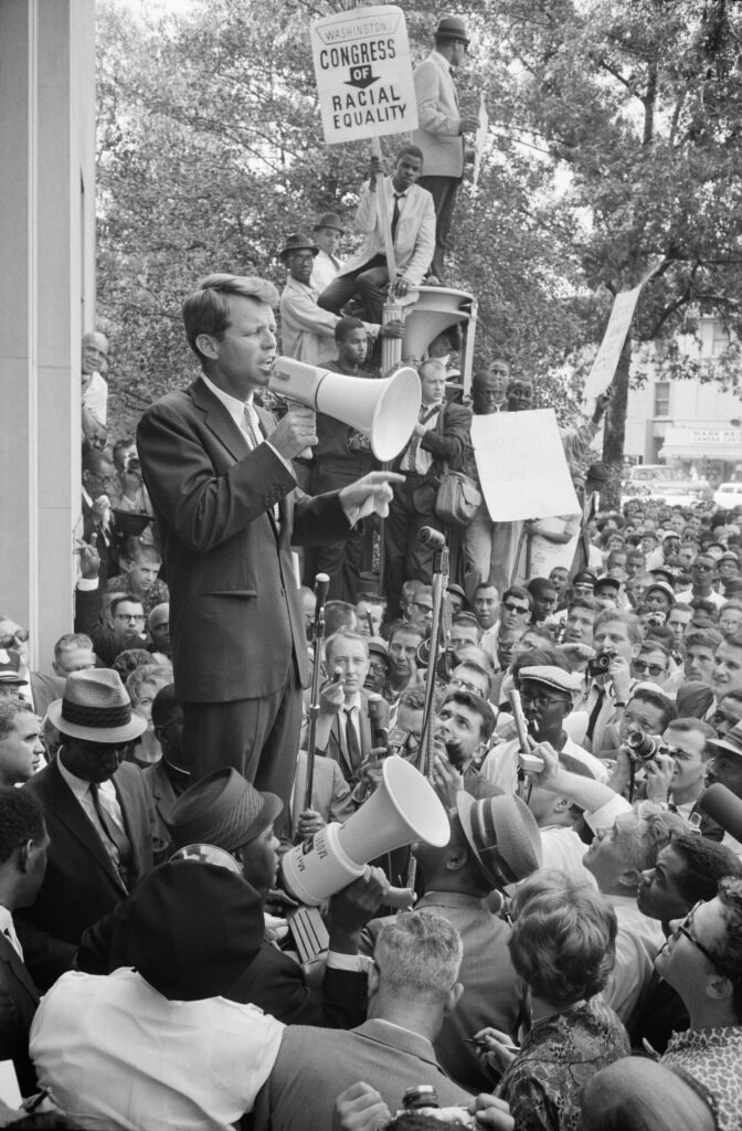 Attorney General Robert F. Kennedy is delivering a speech to a crowd of African Americans and whites through a megaphone outside the Justice Department; a sign for the Congress of Racial Equality is prominently displayed.