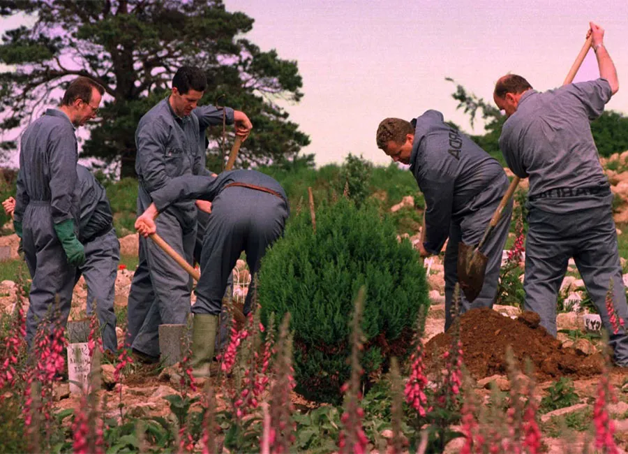 Several Gardaí in grey forensic coveralls dig with shovels at a pet cemetery, surrounded by small headstones, shrubs, and flowering plants, as they carefully excavate the ground during a search operation for Annie McCarrick's body.