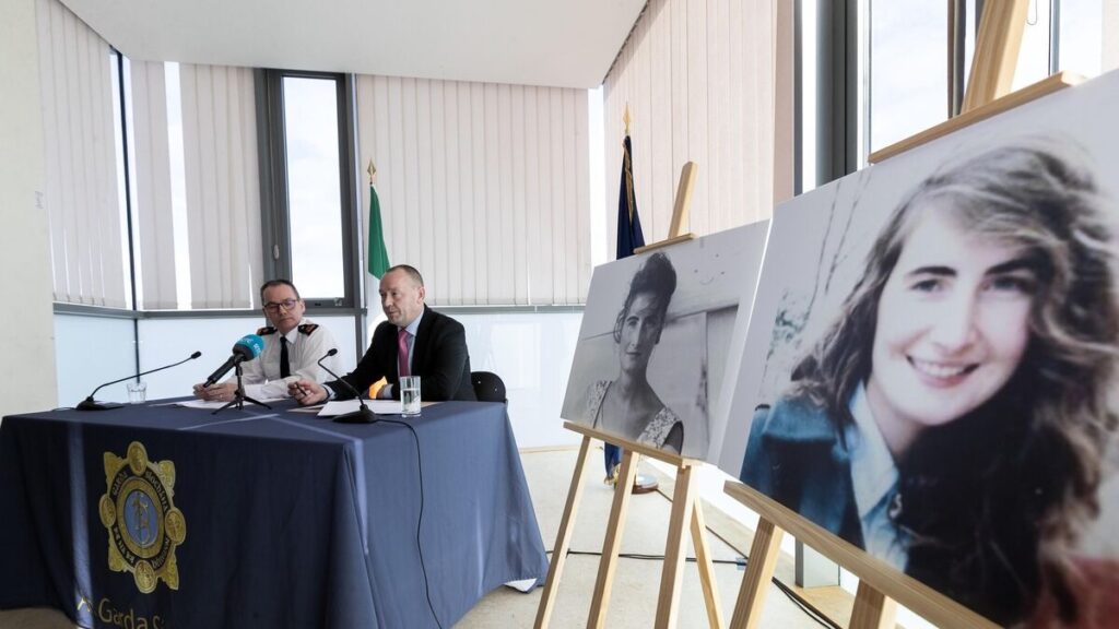 Superintendent Tim Burke and Detective Superintendent Eddie Carroll sit at a press table at Irishtown Garda Station during a media briefing marking the 30th anniversary of Annie McCarrick’s disappearance, with microphones in front of them and large portrait photographs of Annie displayed on easels beside them