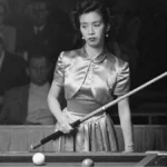 Black-and-white photograph of Japanese billiards champion Masako Katsura standing at a billiards table during a 1952 World Three-Cushion Billiards Tournament appearance in San Francisco. She wears a satin dress, holds a cue diagonally across her body, and looks focused while spectators sit in the dim background.