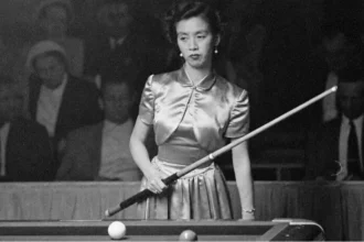 Black-and-white photograph of Japanese billiards champion Masako Katsura standing at a billiards table during a 1952 World Three-Cushion Billiards Tournament appearance in San Francisco. She wears a satin dress, holds a cue diagonally across her body, and looks focused while spectators sit in the dim background.