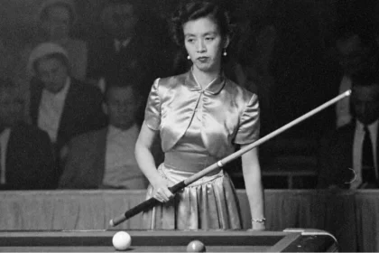 Black-and-white photograph of Japanese billiards champion Masako Katsura standing at a billiards table during a 1952 World Three-Cushion Billiards Tournament appearance in San Francisco. She wears a satin dress, holds a cue diagonally across her body, and looks focused while spectators sit in the dim background.