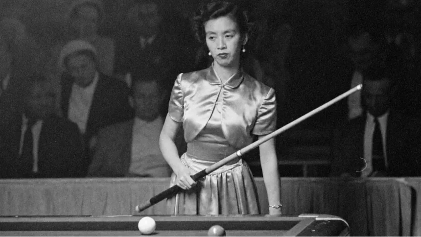 Black-and-white photograph of Japanese billiards champion Masako Katsura standing at a billiards table during a 1952 World Three-Cushion Billiards Tournament appearance in San Francisco. She wears a satin dress, holds a cue diagonally across her body, and looks focused while spectators sit in the dim background.