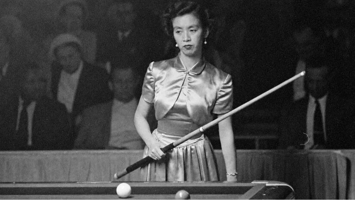Black-and-white photograph of Japanese billiards champion Masako Katsura standing at a billiards table during a 1952 World Three-Cushion Billiards Tournament appearance in San Francisco. She wears a satin dress, holds a cue diagonally across her body, and looks focused while spectators sit in the dim background.