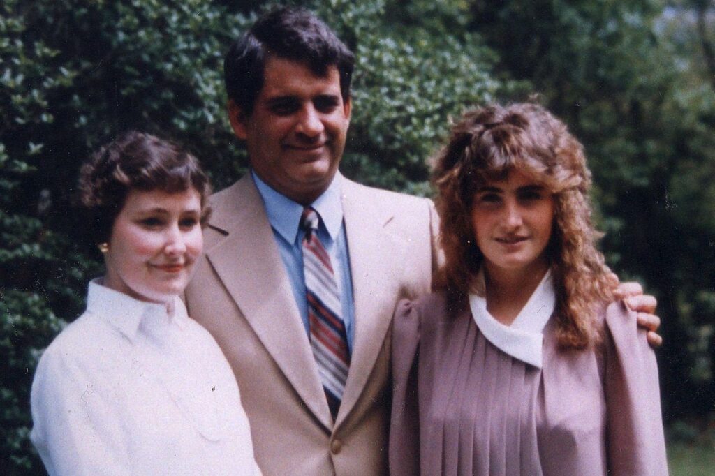 Family photo of Annie McCarrick standing between her parents, John and Nancy McCarrick, outdoors; the three stand close together, with her father in a suit and tie and both women in light-coloured outfits, smiling softly in a leafy setting.