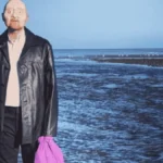 Promotional image for Mr. Bergmann of Deadman’s Point, showing a man in a dark coat standing on a rocky shoreline with the sea behind him and holding a bright purple bag, referencing the unidentified man known as Peter Bergmann.