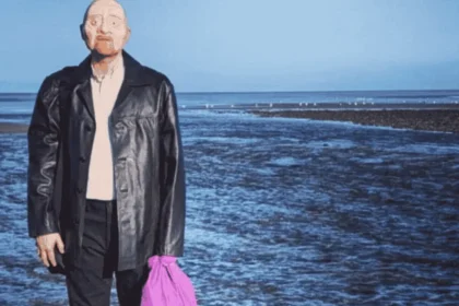 Promotional image for Mr. Bergmann of Deadman’s Point, showing a man in a dark coat standing on a rocky shoreline with the sea behind him and holding a bright purple bag, referencing the unidentified man known as Peter Bergmann.