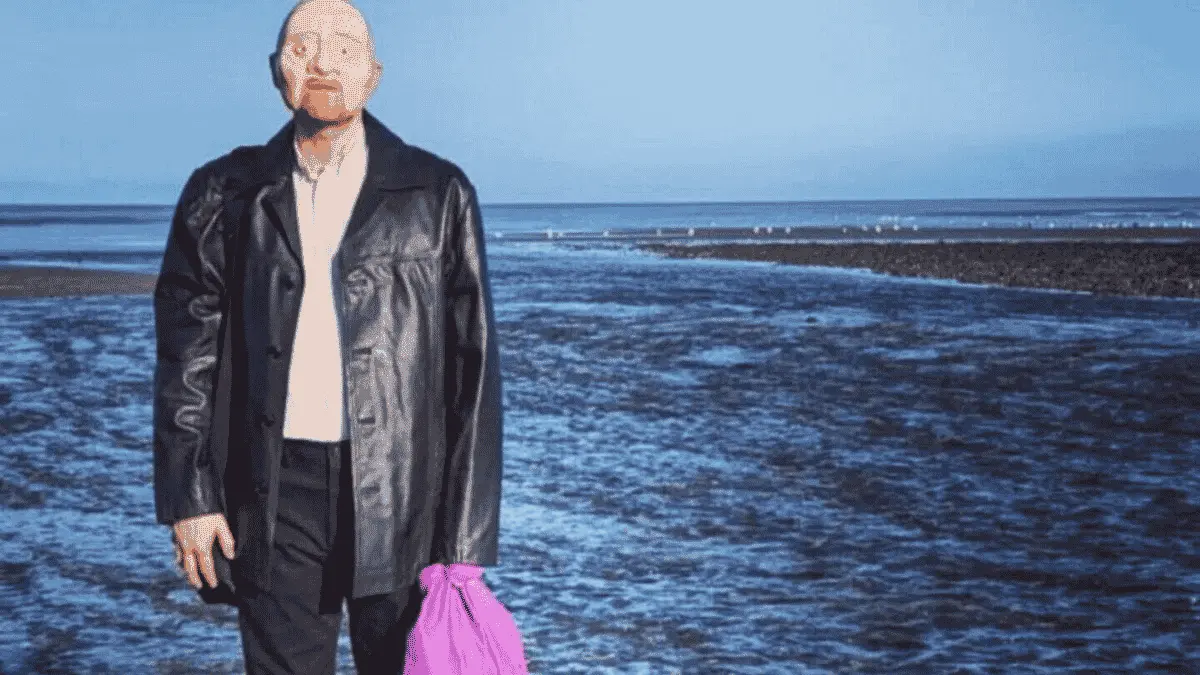 Promotional image for Mr. Bergmann of Deadman’s Point, showing a man in a dark coat standing on a rocky shoreline with the sea behind him and holding a bright purple bag, referencing the unidentified man known as Peter Bergmann.