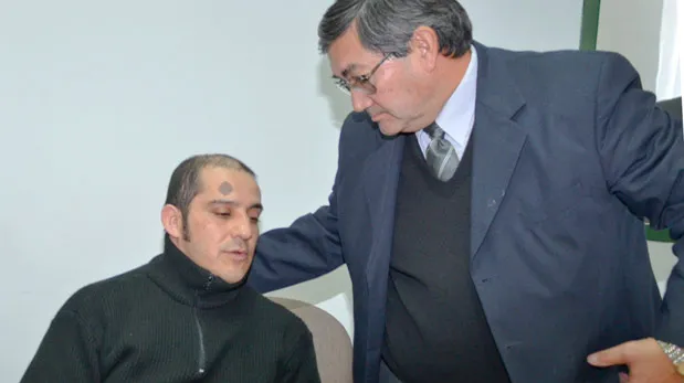 Marcos Gabriel “El Tosco” Díaz sits in court with a visible forehead tattoo while an older man in a suit, his lawyer, stands beside him with a hand on his shoulder.