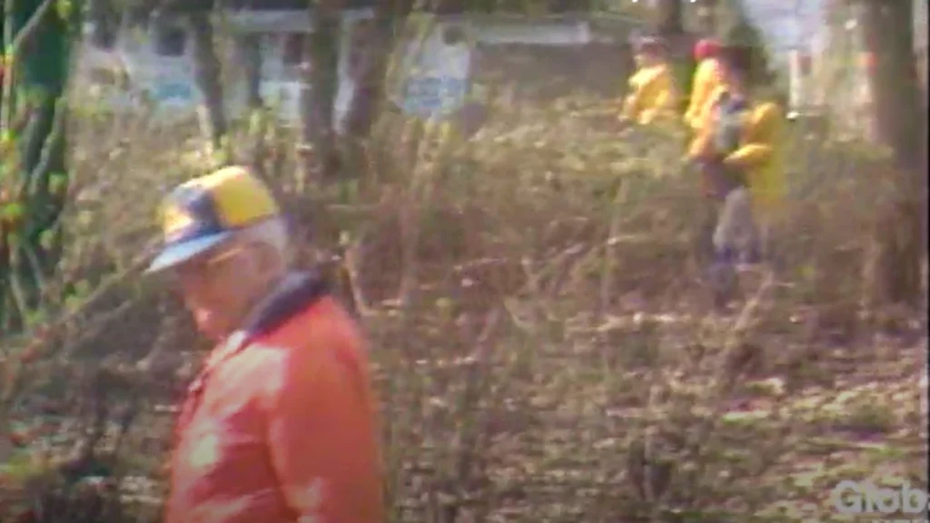 Searchers in orange and yellow safety gear walk shoulder to shoulder across an outdoor area while conducting a grid search for missing 10-year-old Jo-Anne Pedersen.