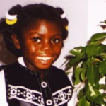 Victoria Climbié smiling beside a leafy houseplant, wearing a black-and-white outfit with yellow-and-white hair ties.