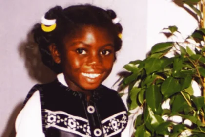 Victoria Climbié smiling beside a leafy houseplant, wearing a black-and-white outfit with yellow-and-white hair ties.