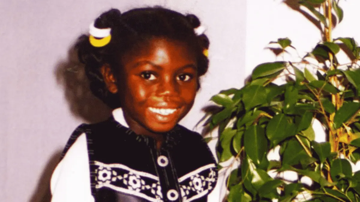 Victoria Climbié smiling beside a leafy houseplant, wearing a black-and-white outfit with yellow-and-white hair ties.