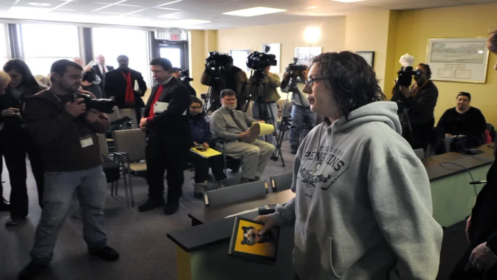 Joy Burkholder, the sister of murder victim Jennifer Daugherty, standing at a podium facing members of the media during a press conference or court recess related to the "Greensburg Six" case.