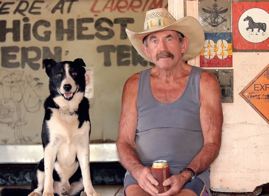 Paddy Moriarty sits at the Larrimah Hotel wearing a cowboy hat and holding a beer, beside his black-and-white dog, Rover.