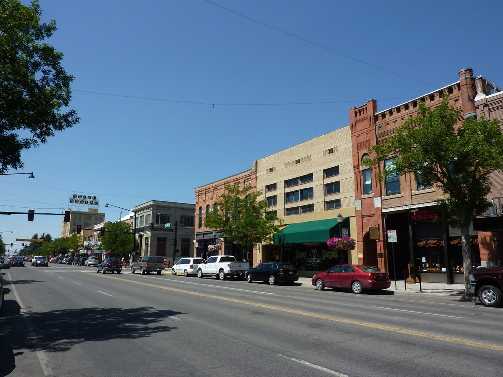 View along Main Street in the historic district, with rows of brick commercial buildings, parked cars, leafy street trees and the Hotel Baxter sign visible in the distance under a clear blue sky.