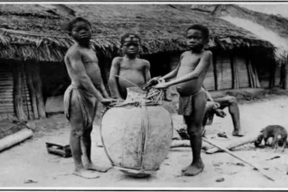 Three Congolese children stand next to a large basket used for transporting agricultural goods or trade items, circa early 1900s.