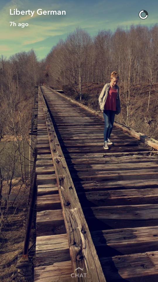 Snapchat photo posted from Liberty German’s account showing a Abby Williams walking across the wooden Monon High Bridge, surrounded by leafless trees under a clear sky.