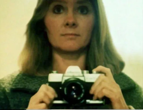 Patricia Meehan takes a mirror self-portrait while holding a camera in front of her, wearing a dark sweater with her long hair loose around her shoulders.