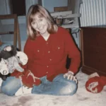 Patricia Meehan sits cross-legged on a carpeted floor, smiling at the camera while holding a large stuffed toy, dressed in a red shirt and blue jeans.