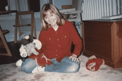 Patricia Meehan sits cross-legged on a carpeted floor, smiling at the camera while holding a large stuffed toy, dressed in a red shirt and blue jeans.