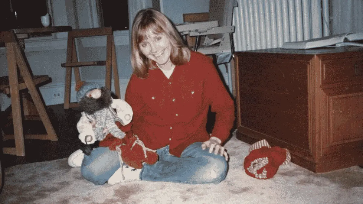 Patricia Meehan sits cross-legged on a carpeted floor, smiling at the camera while holding a large stuffed toy, dressed in a red shirt and blue jeans.
