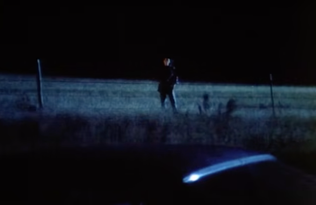 Dramatized scene from Unsolved Mysteries depicting Patricia Meehan standing alone behind a roadside fence at night, illuminated by vehicle headlights after the crash linked to her disappearance.
