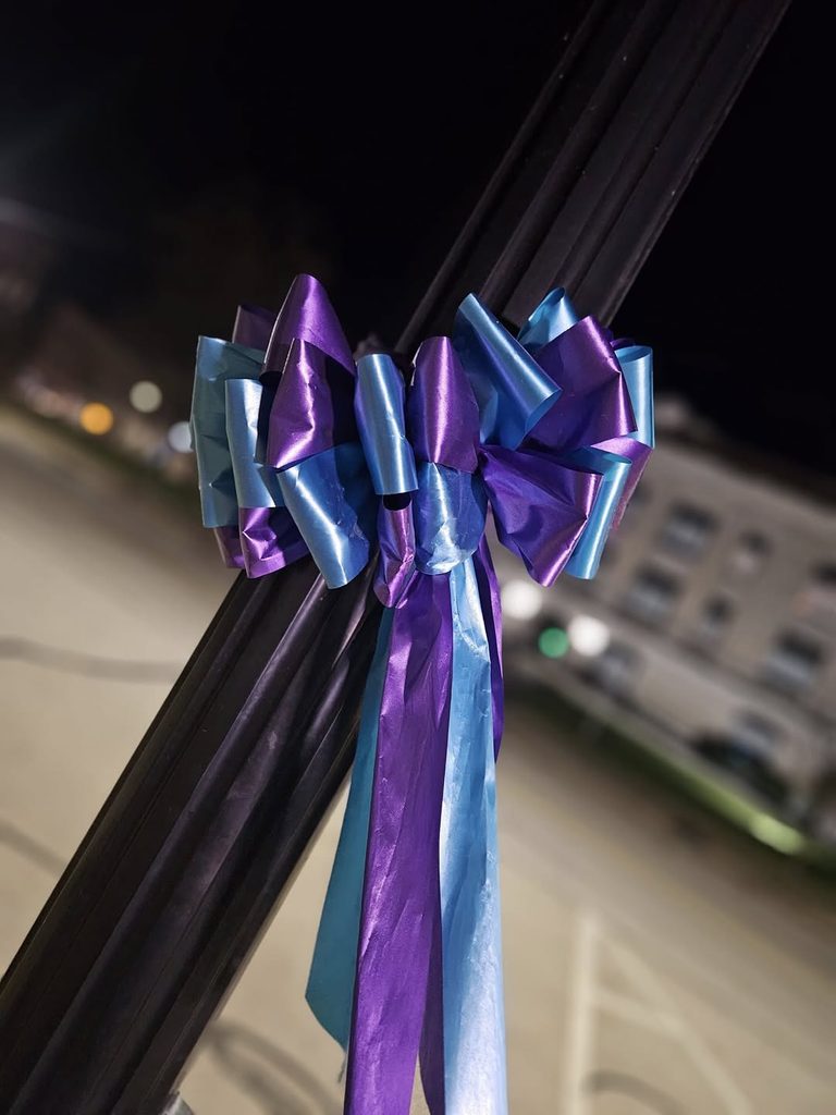 Purple and teal memorial ribbons tied around black metal poles, with a blurred building and lights in the background.