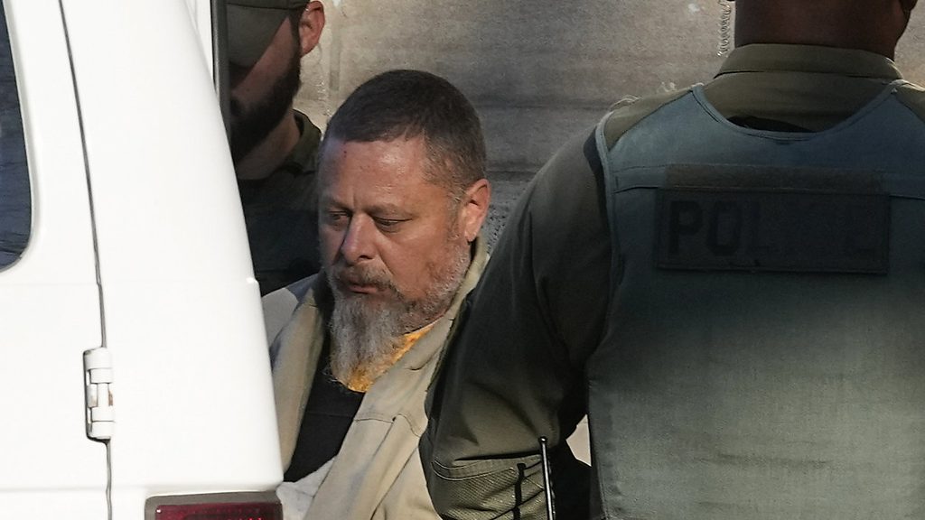 Richard Allen, wearing a tan jacket and sporting a grey beard, is escorted by law enforcement officers beside a white vehicle outside a courthouse.