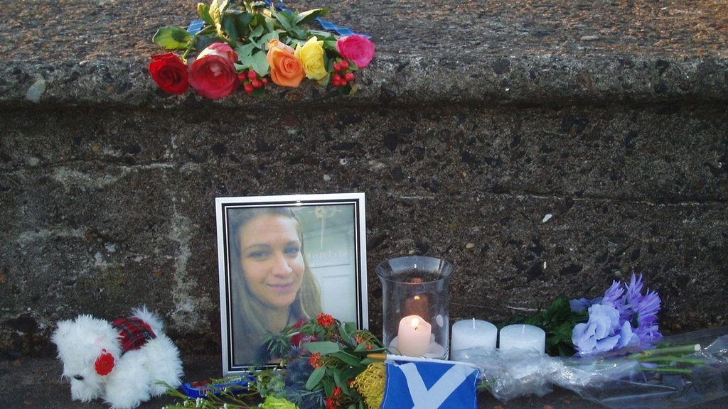Memorial for Annie Börjesson featuring her framed photograph, flowers, lit candles, a white teddy bear and a small Scottish Saltire flag arranged against a stone wall at Prestwick Beach, Scotland