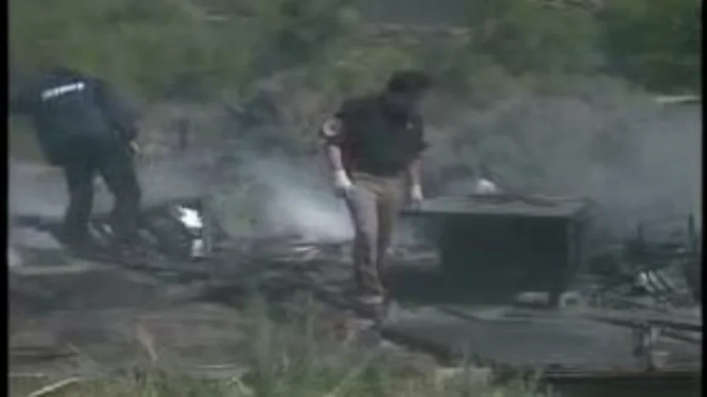 Investigators and law enforcement personnel searching through the smoking, charred ruins of a burned trailer