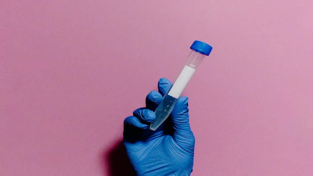 Blue gloved hand holds a test tube against a pastel pink background. 