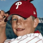 A young Garrett Bardsley smiles and flexes his arm while wearing a red Philadelphia Phillies baseball cap and a white striped shirt, with mountains in the background.