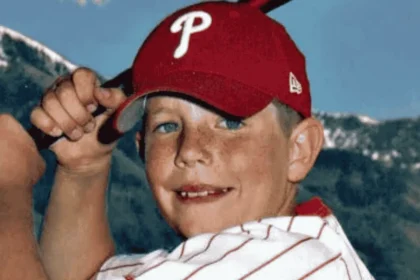 A young Garrett Bardsley smiles and flexes his arm while wearing a red Philadelphia Phillies baseball cap and a white striped shirt, with mountains in the background.