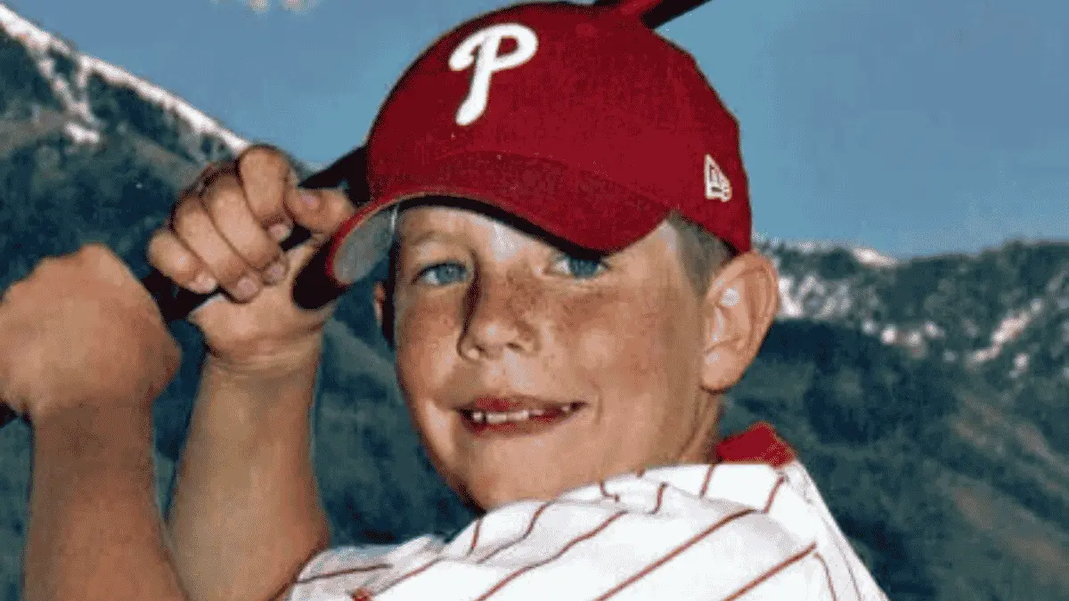 A young Garrett Bardsley smiles and flexes his arm while wearing a red Philadelphia Phillies baseball cap and a white striped shirt, with mountains in the background.
