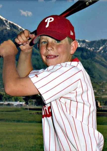 Garrett Bardsley smiling in a Philadelphia Phillies baseball jersey and red cap, holding a bat in a batting stance outdoors with mountains in the background.