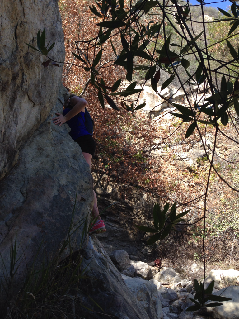 A hiker climbs a large rock near Tangerine Falls while an injured red-haired woman is barely visible among the rocks in the background