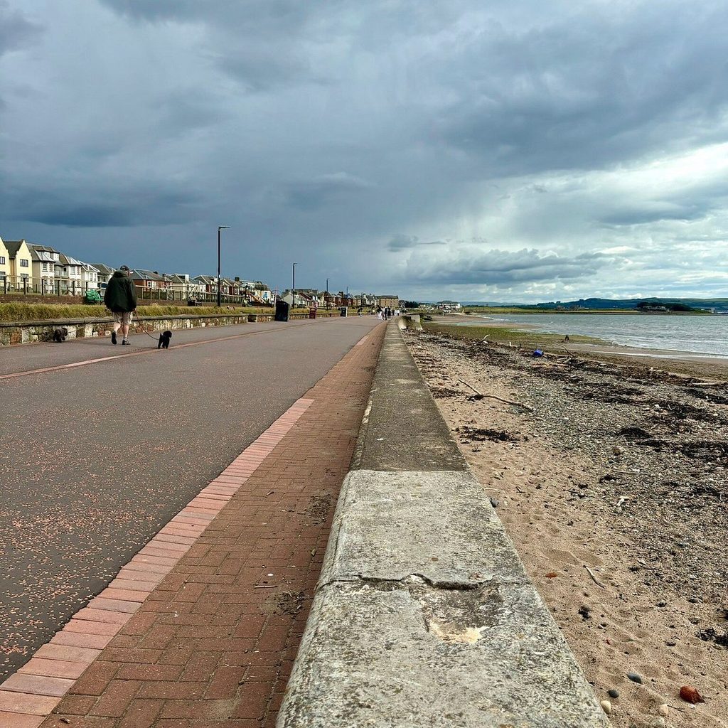 View of Prestwick Beach in Ayrshire, Scotland, showing a seaside promenade, concrete seawall, sandy shoreline and people walking dogs beneath dark, cloudy skies. Annie Börjesson's body was discovered on this beach on December 4, 2005.
