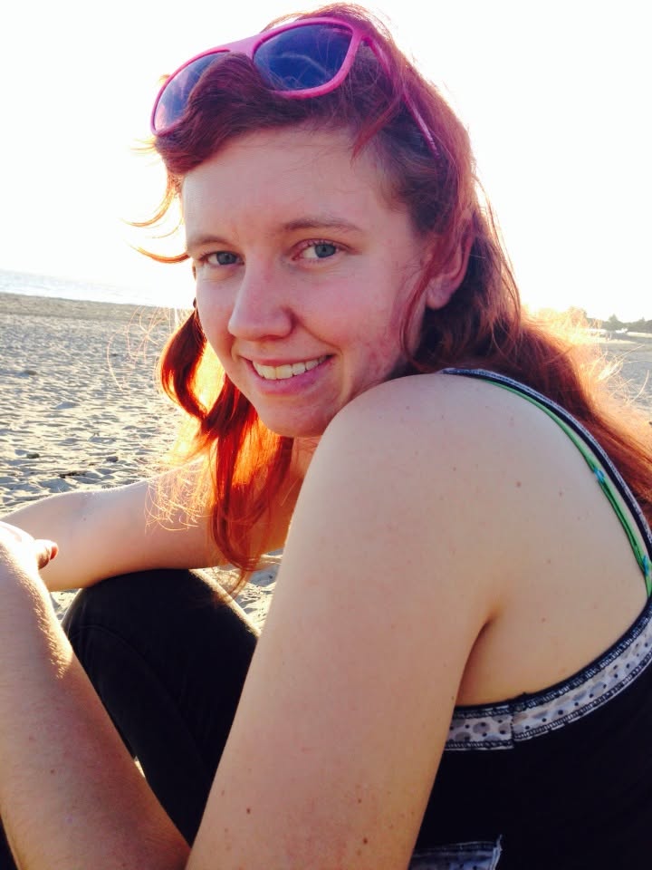 Portrait of Saylor Guilliams smiling on a beach, with red hair and pink sunglasses resting on her head.