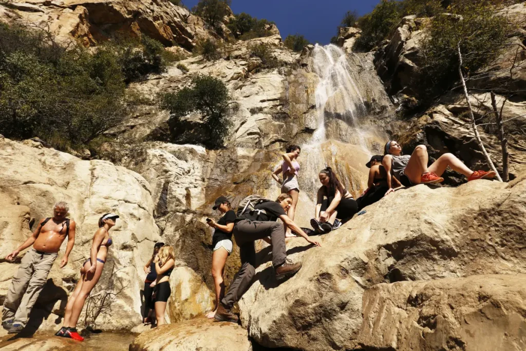 Hikers climb and rest on large rocks beneath Tangerine Falls in Montecito, California, as water cascades down the rocky cliff.