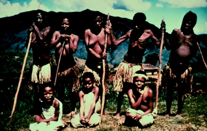 Five women with advanced kuru standing with walking sticks and three girls seated in Papua New Guinea in 1957.